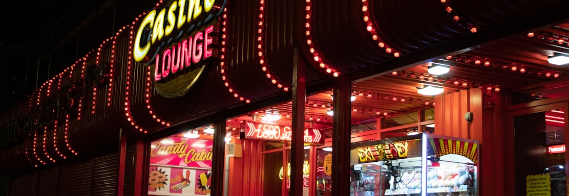 A brightly lit casino lounge entrance adorned with neon lights. The exterior features colorful signage including the words 'Casino' and 'Lounge' in vivid colors, with additional lights framing the building. Inside, a glimpse of a claw machine filled with plush toys and arcade games is visible. The area exudes an inviting yet bustling atmosphere typical of entertainment venues.